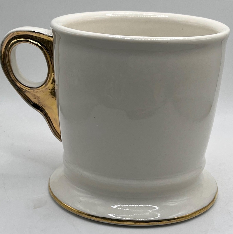 May include: White ceramic shaving mug with a gold-trimmed handle and base. The mug has a wide, round opening and a slightly flared base.