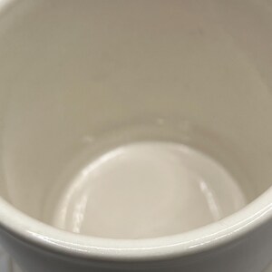 May include: White ceramic mug with a gold design on the bottom. The mug is empty and the inside is visible.