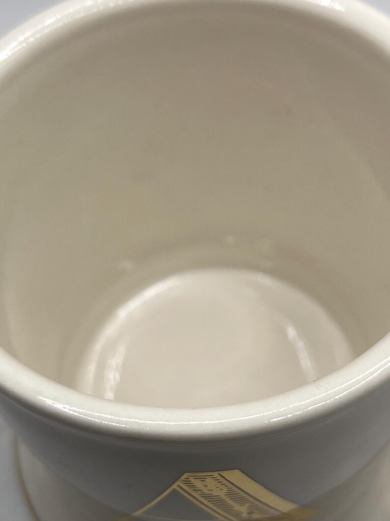 May include: White ceramic mug with a gold design on the bottom. The mug is empty and the inside is visible.