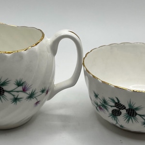 Puede incluir: Un juego de jarra de crema y azucarero de porcelana blanca con ribete dorado y un diseño de piñas verdes y negras.