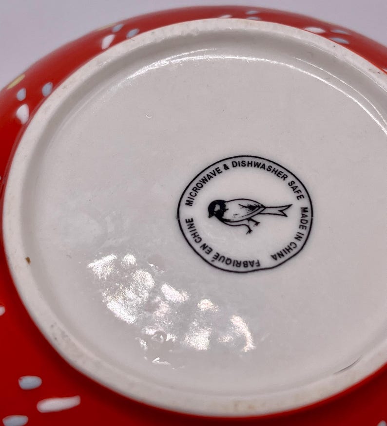 Puede incluir: Un cuenco de cer&aacute;mica rojo con lunares blancos y azules. La base del cuenco es blanca con un sello circular negro que dice "MICROONDAS Y LAVAVAJILLAS" y "HECHO EN CHINA". Una ilustraci&oacute;n de un p&aacute;jaro est&aacute; en el centro.