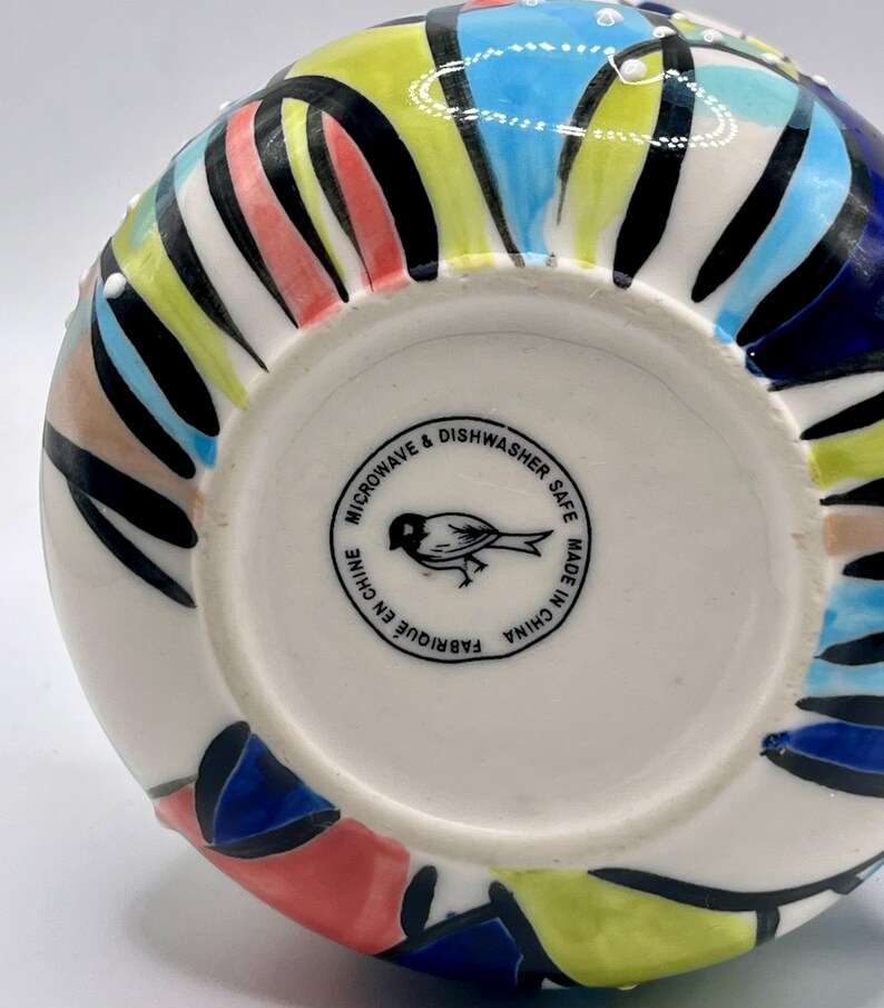 Może przedstawiać: Ceramiczny wazon z kolorowym, abstrakcyjnym wzorem. Wazon ma białą podstawę z wzorem niebieskich, ż&oacute;łtych, czerwonych i czarnych kształt&oacute;w. Na spodzie wazonu znajduje się napis "Microwave & Dishwasher Safe Made in China".