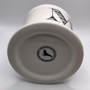 Puede incluir: Taza de cer&aacute;mica blanca con un dise&ntilde;o de letra "N" negra. La taza tiene una base ancha y una parte superior ligeramente acampanada. La parte inferior de la taza tiene un sello circular con un p&aacute;jaro y el texto "Dishwasher & Microwave Safe".