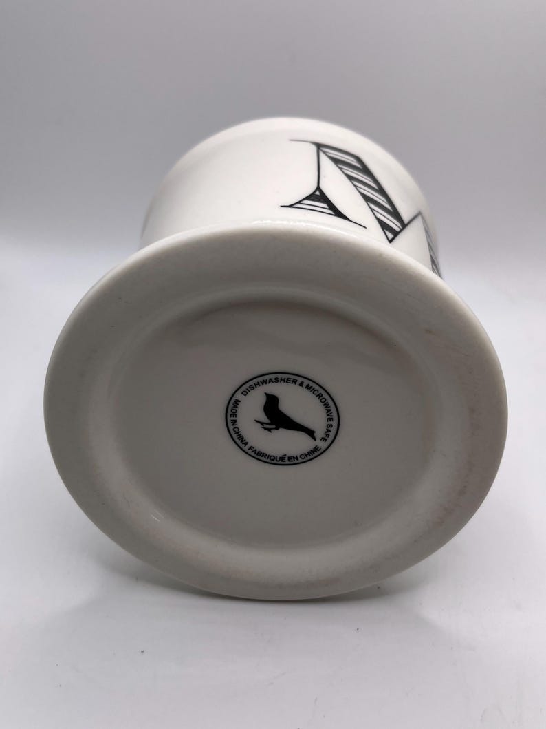Puede incluir: Taza de cer&aacute;mica blanca con un dise&ntilde;o de letra "N" negra. La taza tiene una base ancha y una parte superior ligeramente acampanada. La parte inferior de la taza tiene un sello circular con un p&aacute;jaro y el texto "Dishwasher & Microwave Safe".