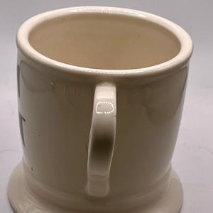 May include: Cream-colored ceramic mug with a rounded base and a sturdy handle. The mug has a wide opening and a smooth, glossy finish. The handle is curved and attached to the side of the mug. The mug is designed for hot beverages.