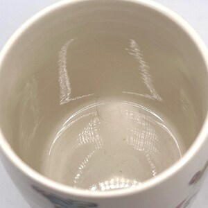May include: A cream-coloured ceramic mug with a wide opening. The interior is smooth and light-toned, with subtle colour variations. The mug's exterior features a faint floral design. Suitable for hot or cold drinks.