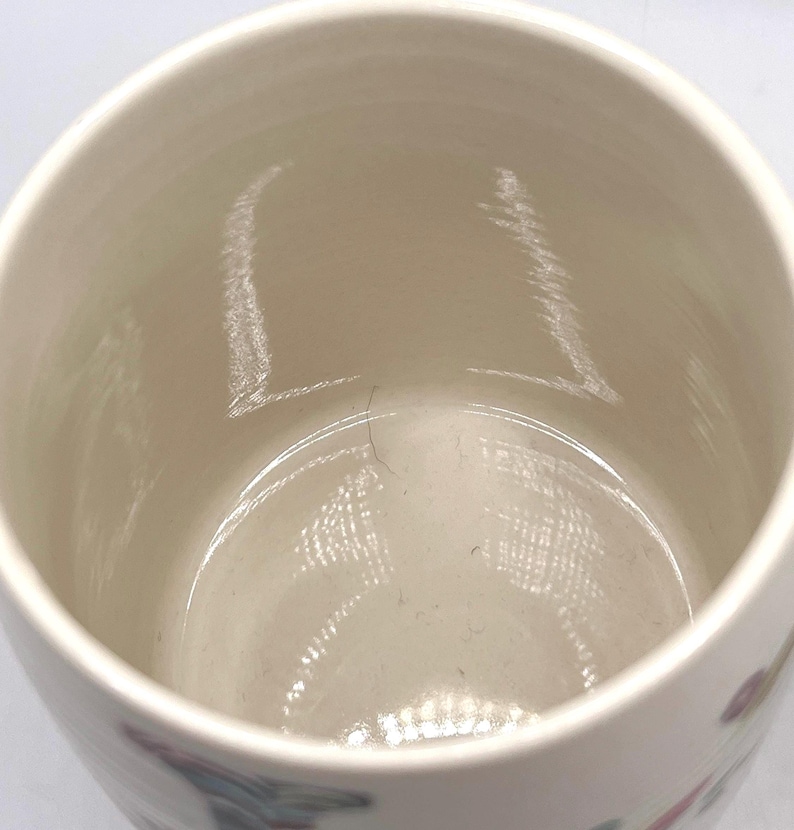 May include: A cream-coloured ceramic mug with a wide opening. The interior is smooth and light-toned, with subtle colour variations. The mug's exterior features a faint floral design. Suitable for hot or cold drinks.