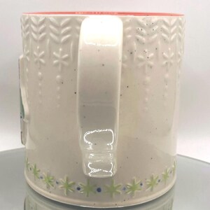 May include: Cream-colored ceramic mug with a red interior and a white handle. The mug features a repeating pattern of embossed snowflakes and leaves, with green and blue star-shaped designs around the base. The mug is speckled with small black dots.
