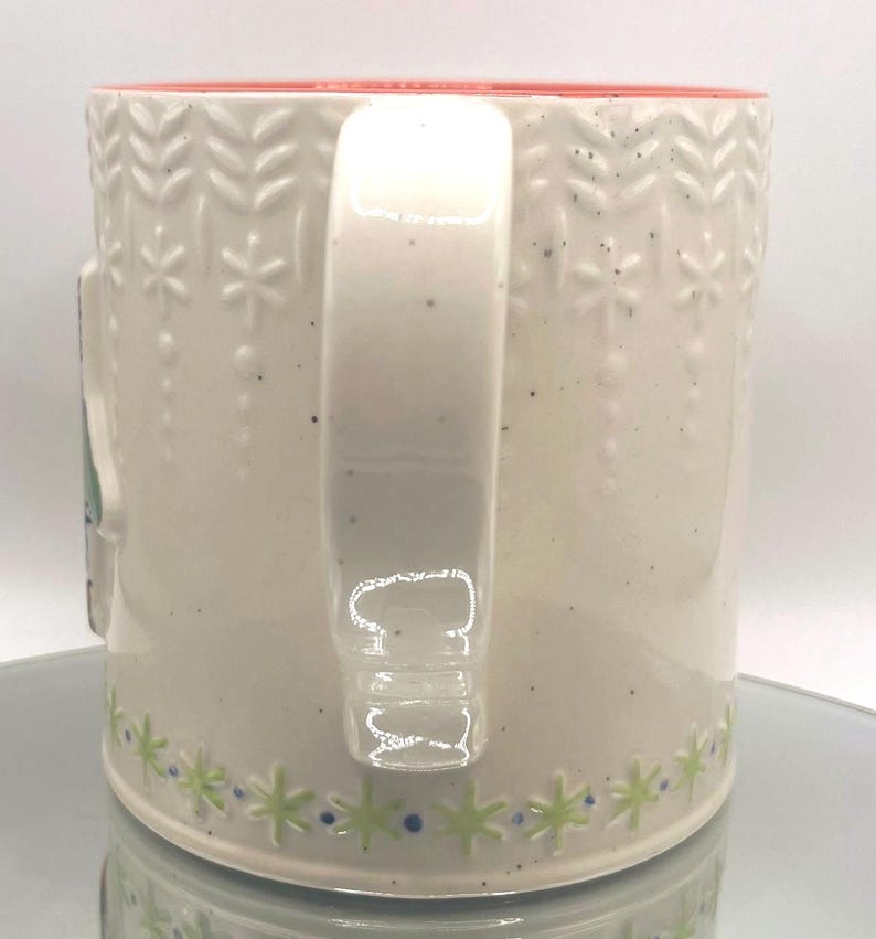 May include: Cream-colored ceramic mug with a red interior and a white handle. The mug features a repeating pattern of embossed snowflakes and leaves, with green and blue star-shaped designs around the base. The mug is speckled with small black dots.