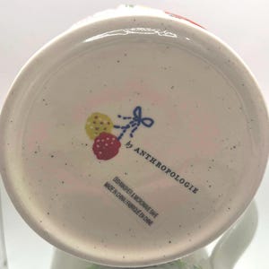 May include: White ceramic mug base with a speckled finish. The bottom features a design of a yellow and red fruit with a blue bow, and the text "by ANTHROPOLOGIE". The text "DISHWASHER & MICROWAVE SAFE" is also visible.
