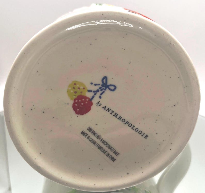 May include: White ceramic mug base with a speckled finish. The bottom features a design of a yellow and red fruit with a blue bow, and the text "by ANTHROPOLOGIE". The text "DISHWASHER & MICROWAVE SAFE" is also visible.