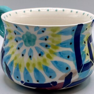 May include: A ceramic mug with a turquoise handle and a white interior. The exterior is decorated with a colorful floral pattern in shades of blue, green, and yellow, with black dot accents. The mug has a wide opening.