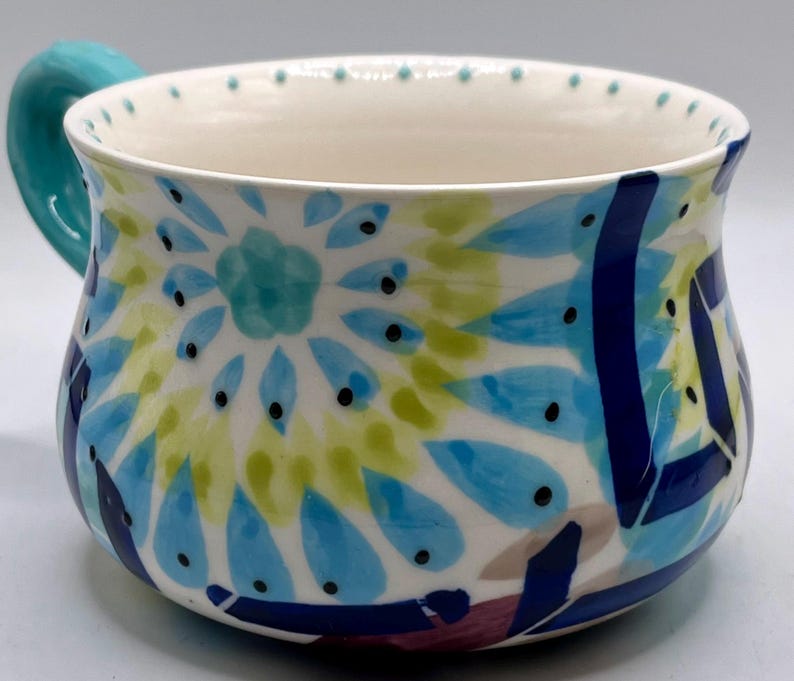 May include: A ceramic mug with a turquoise handle and a white interior. The exterior is decorated with a colorful floral pattern in shades of blue, green, and yellow, with black dot accents. The mug has a wide opening.
