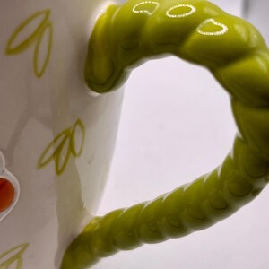 May include: White ceramic mug with a bright green, textured handle. The mug features yellow leaf designs and colorful floral accents. The handle is a unique, twisted design, adding a whimsical touch to the drinkware.