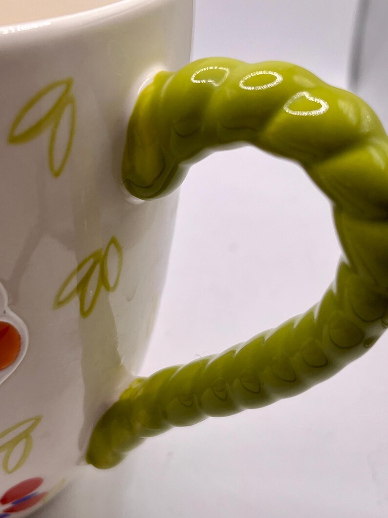 May include: White ceramic mug with a bright green, textured handle. The mug features yellow leaf designs and colorful floral accents. The handle is a unique, twisted design, adding a whimsical touch to the drinkware.