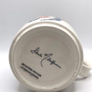 May include: Cream ceramic mug with a handle. The base of the mug features a signature and the text "Dishwasher & Microwave Safe Made in China / Fabriqu&eacute; en Chine". The mug has a white interior.