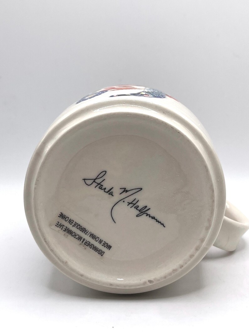 May include: Cream ceramic mug with a handle. The base of the mug features a signature and the text "Dishwasher & Microwave Safe Made in China / Fabriqu&eacute; en Chine". The mug has a white interior.