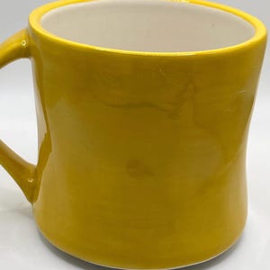 Puede incluir: Una taza de cer&aacute;mica de color amarillo mostaza con un asa curvada. El interior es blanco, contrastando con el acabado brillante del exterior. La taza tiene un dise&ntilde;o simple y redondeado, adecuada para bebidas calientes.