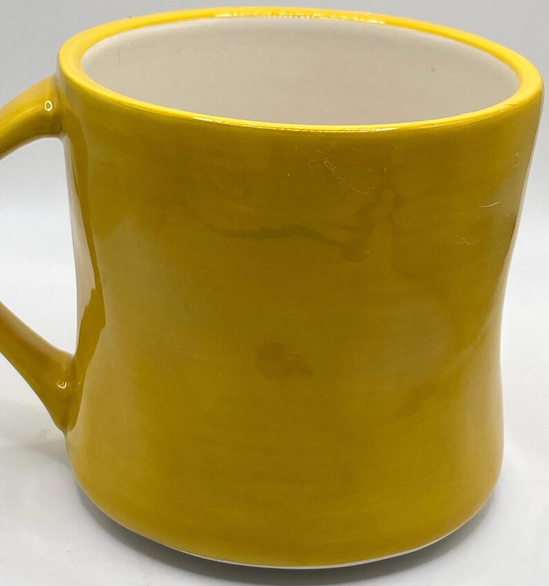 Puede incluir: Una taza de cer&aacute;mica de color amarillo mostaza con un asa curvada. El interior es blanco, contrastando con el acabado brillante del exterior. La taza tiene un dise&ntilde;o simple y redondeado, adecuada para bebidas calientes.