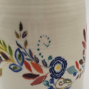 May include: White ceramic mug with a hand-painted floral design. The design features blue, green, red, and brown flowers and leaves.