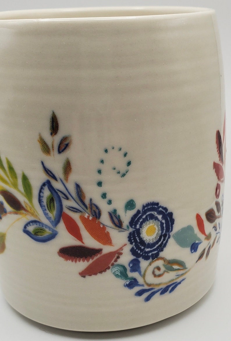 May include: White ceramic mug with a hand-painted floral design. The design features blue, green, red, and brown flowers and leaves.
