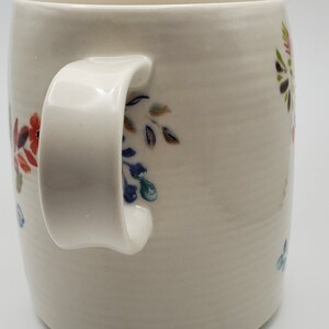 May include: White ceramic mug with a floral design in shades of blue, green, red, and brown. The mug has a rounded handle.