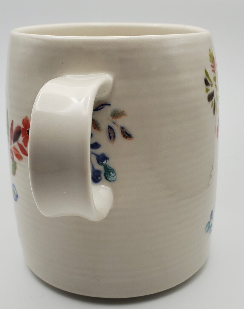 May include: White ceramic mug with a floral design in shades of blue, green, red, and brown. The mug has a rounded handle.