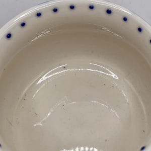 Może przedstawiać: Biała ceramiczna miska z błyszczącym wnętrzem. Obręcz jest ozdobiona r&oacute;wnomiernie rozmieszczonymi niebieskimi kropkami. Wnętrze miski ma subtelne ślady. Miska jest prawdopodobnie używana do serwowania jedzenia lub jako element dekoracyjny.