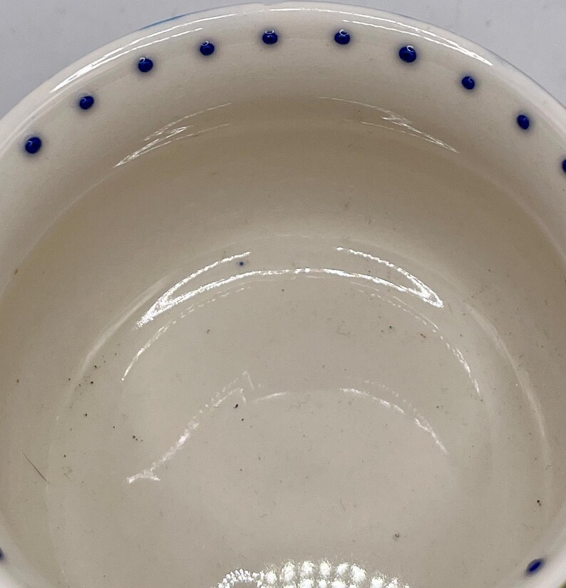 Może przedstawiać: Biała ceramiczna miska z błyszczącym wnętrzem. Obręcz jest ozdobiona r&oacute;wnomiernie rozmieszczonymi niebieskimi kropkami. Wnętrze miski ma subtelne ślady. Miska jest prawdopodobnie używana do serwowania jedzenia lub jako element dekoracyjny.