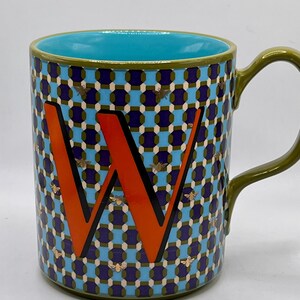 May include: A ceramic mug with a teal interior and a green handle. The exterior features a geometric pattern in blue, purple, and white, with an orange letter "W" and gold bee accents. The mug is designed for beverages.
