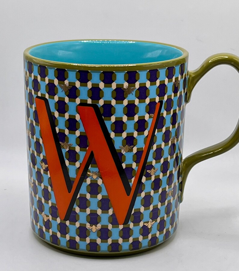 May include: A ceramic mug with a teal interior and a green handle. The exterior features a geometric pattern in blue, purple, and white, with an orange letter "W" and gold bee accents. The mug is designed for beverages.