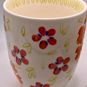 May include: A white ceramic mug with a floral design. The mug features red flowers with blue centers and green leaves. The rim has a yellow dotted pattern. The mug is approximately 4 inches tall.