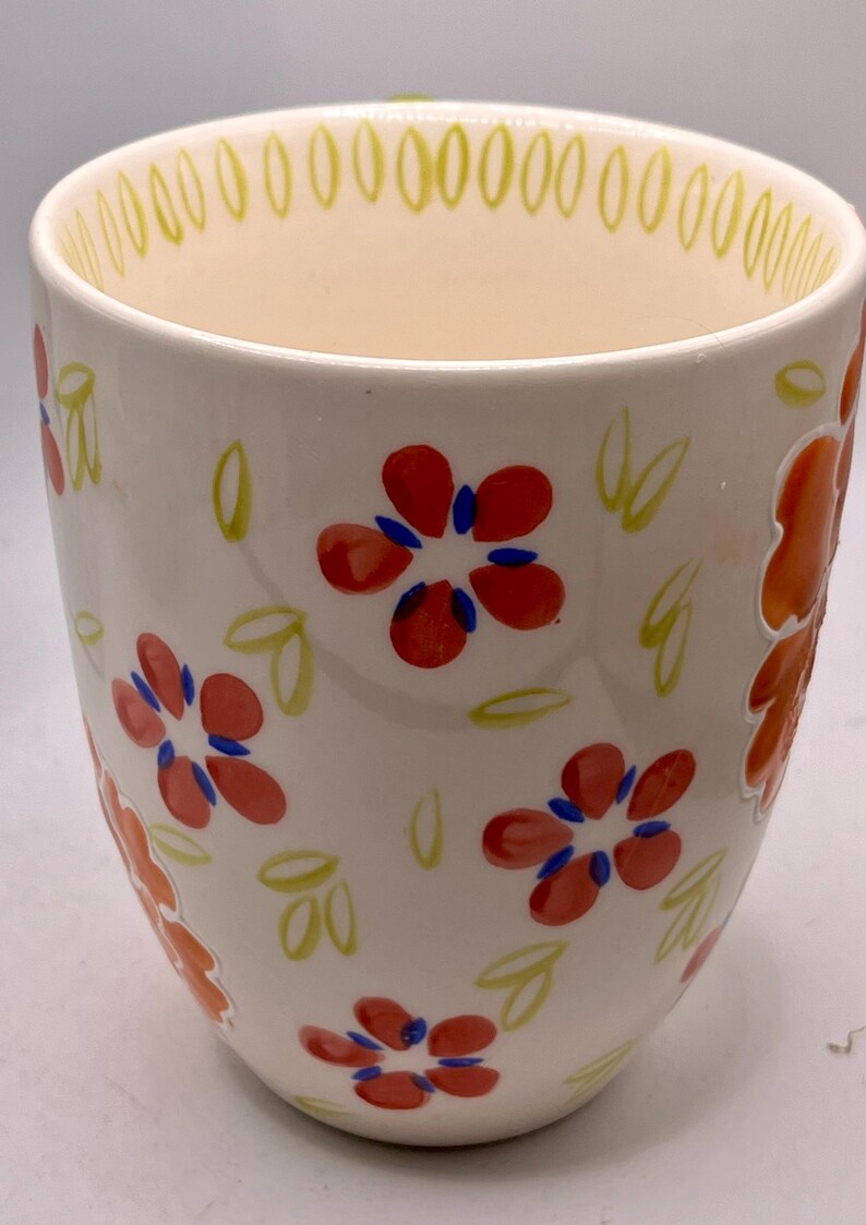 May include: A white ceramic mug with a floral design. The mug features red flowers with blue centers and green leaves. The rim has a yellow dotted pattern. The mug is approximately 4 inches tall.