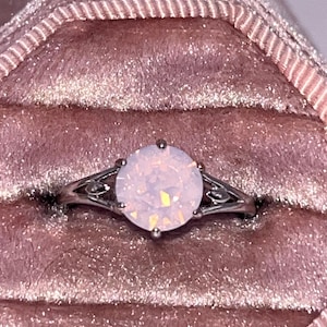 May include: A silver ring with a large, round, opalescent gemstone. The ring is set in a delicate, filigree design and is displayed in a pink velvet ring box. The gemstone has a soft, iridescent glow, reflecting shades of pink and white.