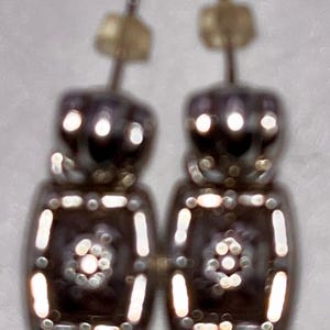 May include: A pair of silver-coloured earrings with a geometric design. Each earring has a rectangular shape with small, round accents and a central floral pattern. The earrings have a post-style backing.