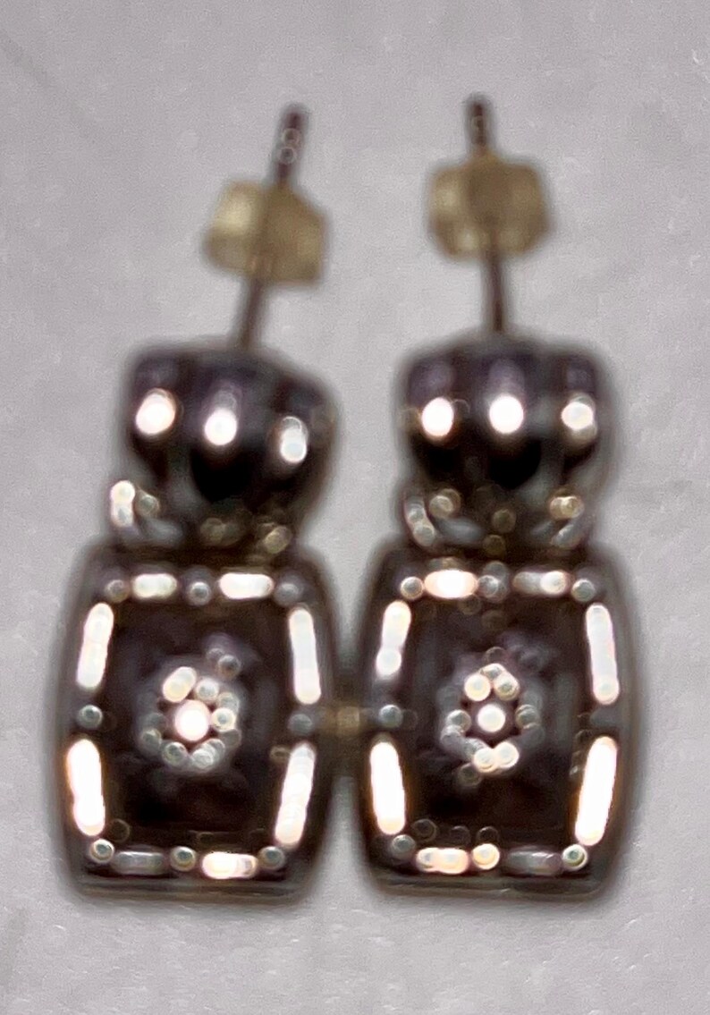May include: A pair of silver-coloured earrings with a geometric design. Each earring has a rectangular shape with small, round accents and a central floral pattern. The earrings have a post-style backing.