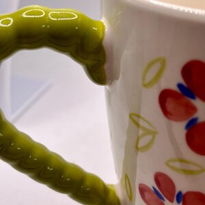 May include: White ceramic mug with a green, textured handle. The mug is decorated with red flowers and green leaves. Yellow lines adorn the rim. A charming and colorful addition to any kitchen.