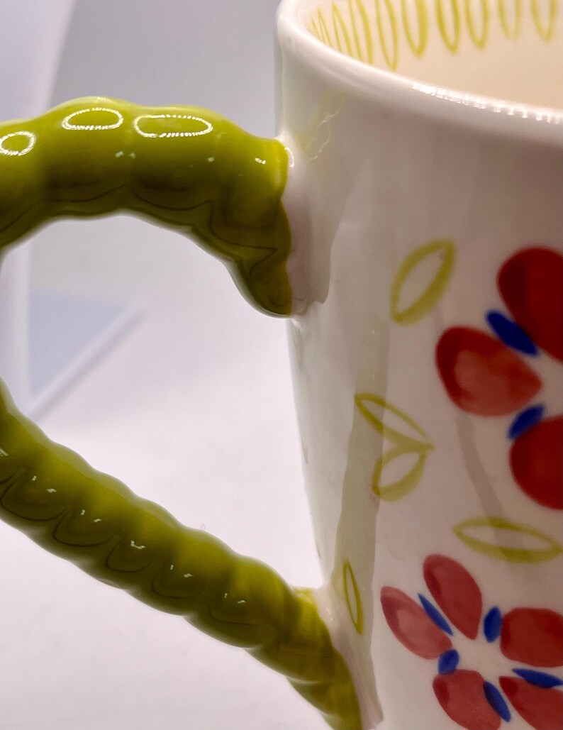 May include: White ceramic mug with a green, textured handle. The mug is decorated with red flowers and green leaves. Yellow lines adorn the rim. A charming and colorful addition to any kitchen.