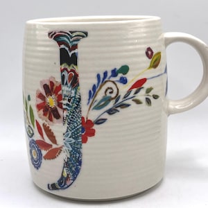 Könnte beinhalten: Cremefarbene Keramiktasse mit einem großen, farbenfrohen floralen "J"-Design. Der Buchstabe ist mit einer Mischung aus Mustern und Farben gefüllt, darunter Blau, Rot und Grün. Die Tasse hat einen gebogenen Griff und eine strukturierte Oberfläche.