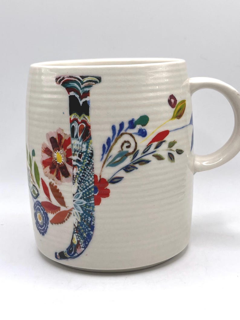May include: Cream ceramic mug featuring a large, colourful floral "J" design. The letter is filled with a mix of patterns and colours, including blue, red, and green. The mug has a curved handle and a textured surface.
