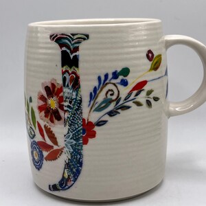 May include: Cream ceramic mug featuring a large, colourful floral "J" design. The letter is filled with a mix of patterns and colours, including red, blue, and green. The mug has a ribbed texture and a curved handle.