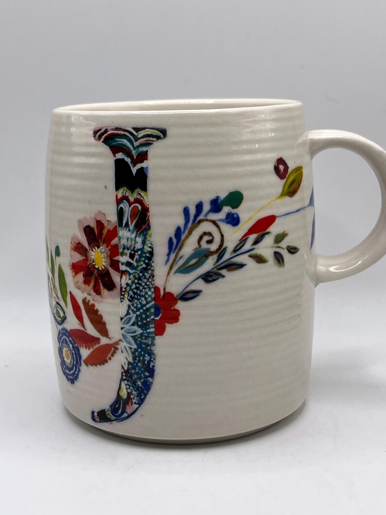May include: Cream ceramic mug featuring a large, colourful floral "J" design. The letter is filled with a mix of patterns and colours, including red, blue, and green. The mug has a ribbed texture and a curved handle.