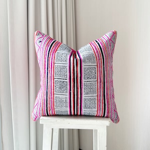 May include: A white pillow with pink and black stripes and geometric patterns. The pillow is sitting on a white stool.