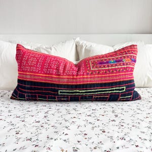 Puede incluir: Un cojín lumbar rectangular que presenta una sección superior roja vibrante con intrincados bordados geométricos blancos y una sección inferior azul marino oscuro con líneas y patrones cosidos en verde lima y naranja.