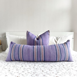 Pode incluir: Uma almofada lombar listrada em roxo e azul com borlas em uma cama branca com uma colcha estampada floral.