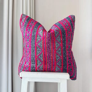 Puede incluir: Una almohada de tiro de color rosa y negro con un diseño geométrico. La almohada está sentada en un taburete blanco.