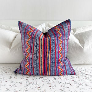 May include: A blue pillow with colorful embroidered stripes and zig-zag patterns. The pillow is on a white bed with a floral patterned bedspread.