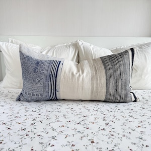 Puede incluir: Un cojín decorativo rectangular con un diseño de patchwork. El cojín presenta secciones de tela azul estampada, tela color crema y tela a rayas grises y negras. El cojín está colocado sobre una cama con ropa de cama blanca y una sábana con estampado floral.