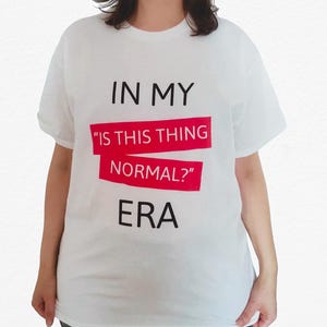 Puede incluir: Camiseta blanca con el texto "IN MY" sobre un rectángulo rojo con el texto "IS THIS THING NORMAL?" y la palabra "ERA" debajo. La camiseta es de estilo casual.