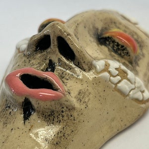 May include: A ceramic mask with a distressed, textured surface. The mask is painted in shades of beige, brown, pink, and white. The mask has a large, open mouth with pink lips and a prominent nose.
